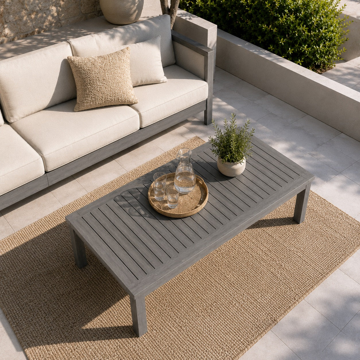 Table basse de jardin en bois gris sur terrasse extérieure avec canapé beige et tapis tressé.